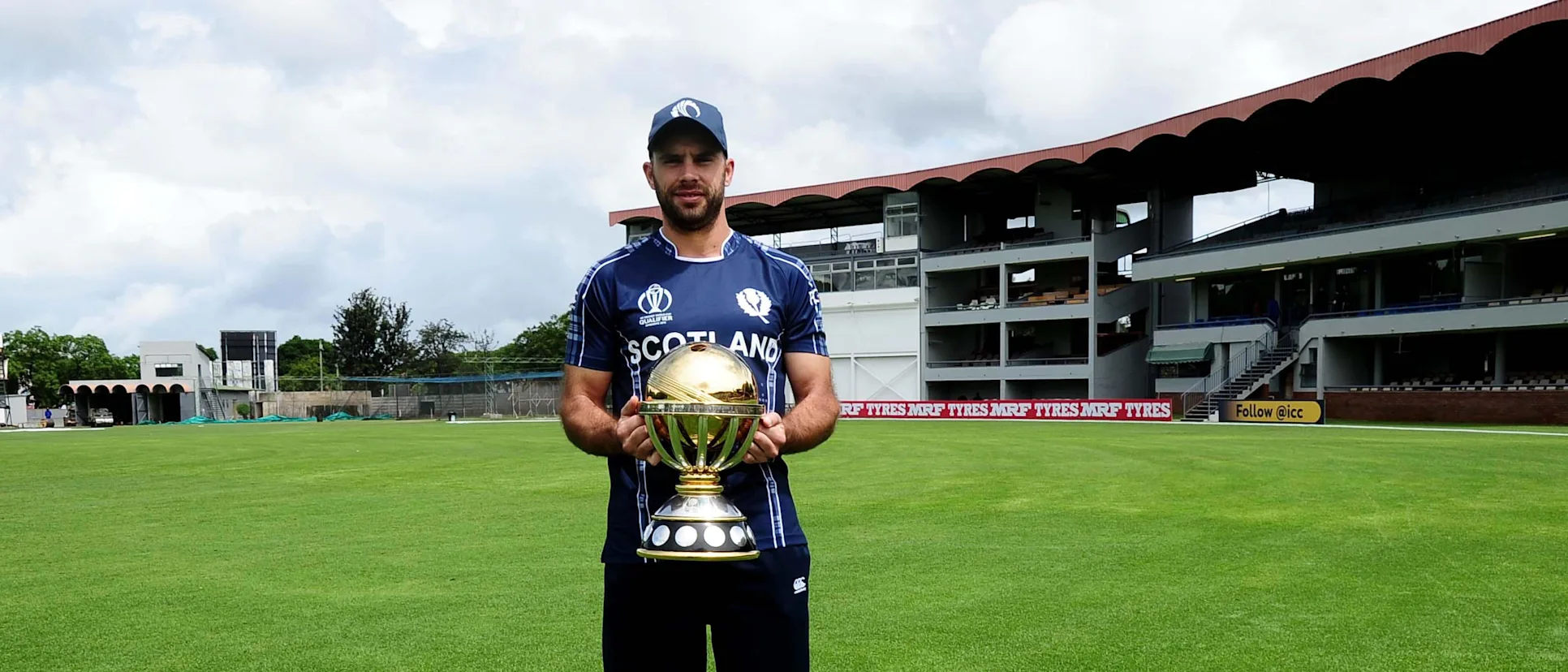 Logo of Scotland Captain Kyle Coetzer holding the ICC CWCQ Trophy at Queens Sports Club.